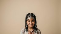 Young Black female child with braided hairstyle adorned with beads, wearing casual clothing against neutral beige background. Portrait suitable for diversity, education, and lifestyle content.