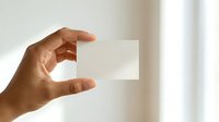 Hand of a person with medium brown skin tone holding a blank white business card or paper against a blurred neutral background, suitable for branding and corporate use.