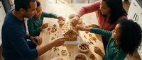 Young multiethnic family with two children gathered around gingerbread house during holiday celebration, sharing festive baking activity together at wooden table with ingredients and decorations.