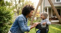 African American mother pushing happy toddler son on backyard swing set during golden hour sunset creating joyful family bonding moment in residential neighborhood.