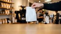 Person with pale skin holding white paper bag in coffee shop with warm lighting and blurred staff background, retail and service concept.