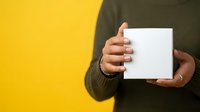 Person with medium skin tone wearing olive green sweater holding blank white box against yellow background, suitable for product packaging mockups and promotional campaigns.