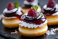 Mini chocolate glazed donuts topped with whipped cream, fresh raspberry and mint leaf garnish on dark slate surface. Sweet pastry dessert closeup.