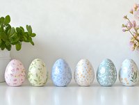 Decorative Easter eggs in pastel colors with delicate floral patterns arranged in row on white surface. Green plant and pink flowers complete spring composition.