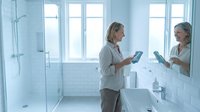 Young blonde woman in casual shirt using smartphone while standing in modern white bathroom with large mirror and glass shower enclosure.