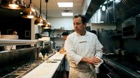 Professional middle-aged Caucasian male chef in white uniform standing in modern commercial restaurant kitchen with stainless steel equipment and copper pendant lights.