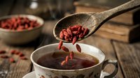 Dried goji berries falling from wooden spoon into herbal tea cup on rustic kitchen table for healthy lifestyle and natural wellness nutrition.