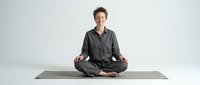 Middle-aged Caucasian woman in gray pajamas practicing meditation in lotus position on yoga mat against white background for wellness and mindfulness content.