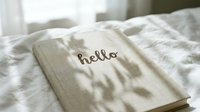 Linen notebook with hello lettering resting on white bed sheets in soft morning light with delicate plant shadows.