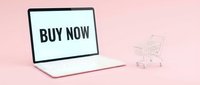 White laptop displaying buy now text beside a miniature shopping cart on a pink background, symbolizing online shopping and e-commerce.