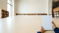 Young professional with dark hair and medium skin tone holding blank white notebook in modern library or office setting with wooden shelves and natural light.