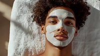 Young African American man with curly hair wearing facial clay mask during skincare routine, looking at camera with confident expression against neutral background.