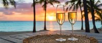 Two champagne glasses on wicker placemat at tropical beach sunset with palm trees and turquoise ocean for romantic celebration and luxury vacation concept.