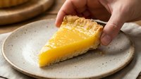 Hand holding slice of homemade lemon tart with golden custard filling and crumbly crust on speckled ceramic plate for dessert presentation and food styling.