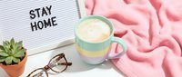 Cozy stay home flat lay with pastel striped mug of latte, pink blanket, succulent plant, glasses, and letter board on white surface.