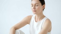 Young adult woman with fair complexion and brown hair, wearing white sleeveless tank top, posing with eyes closed in contemplative pose against neutral background for wellness and lifestyle content.