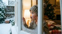 Young woman in knit sweater and beanie looking through frosted window at snowy winter landscape from cozy warm interior with soft lighting.