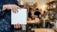 Professional holding blank white card in modern cafe environment with blurred background, suitable for business promotions and marketing materials.