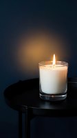 Lit white candle in clear glass container on black table against dark blue background, creating warm ambient glow for relaxation, meditation, home decor, or wellness design projects.