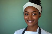Young African American female chef wearing white headband and apron smiling warmly at camera against green background, expressing confidence and professionalism.