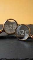 Two brass magnifying glasses with engraved B2B and B2C text on glass lenses, positioned on dark slate surface against warm yellow background for business marketing.