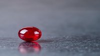 Red translucent pill capsule on dark textured surface with reflection, dramatic lighting and shallow depth of field creates minimalist pharmaceutical composition.