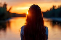 Young woman with long dark hair silhouetted against golden sunset over calm lake. Orange sky reflects on water surface creating peaceful atmospheric scene.