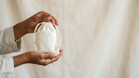 Close-up of hands holding white cotton drawstring pouch against neutral background. Suitable for product packaging, eco-friendly branding, sustainable business concepts.