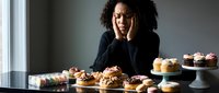 Young African American woman baker feeling overwhelmed surrounded by homemade cupcakes and desserts in modern kitchen. Stress, burnout concept.