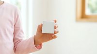 Young adult male with medium skin tone wearing pink long-sleeve shirt holding blank white box in hand against blurred interior background for product mockup and branding purposes.