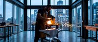 Professional blacksmith working with hot metal and sparks in modern urban workshop with city skyline view through large windows at dusk.
