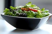 Fresh green salad with cherry tomatoes and sesame seeds served in modern black ceramic bowl on white surface, healthy eating concept for restaurant menu.