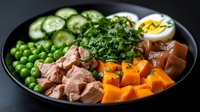 Healthy protein bowl with sliced tuna, boiled egg, green peas, cucumber, sweet potato and fresh herbs on dark plate for balanced nutrition meal.