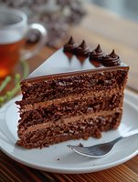 Rich chocolate layer cake slice with creamy ganache filling and decorative piped frosting rosettes on top, served on white plate with tea cup in background.