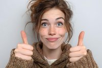 Young woman with freckles showing thumbs up gesture, expressing positivity and approval while wearing cozy brown knit sweater against neutral background.