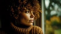 Young African American woman with curly hair in cozy brown sweater looking thoughtfully out window with warm natural lighting for lifestyle content.