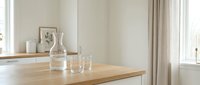 A clear glass bottle and drinking glass sit on a wooden table in a bright, minimalist kitchen with neutral tones, white cabinetry, and natural light from windows.