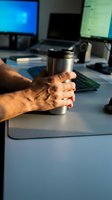 Male hands holding stainless steel travel mug at modern office desk with computer monitors and keyboard in background during work break.