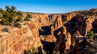 Dramatic red rock canyon landscape with natural stone arches and desert vegetation under clear blue sky, showcasing rugged southwestern terrain formations.