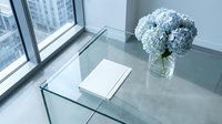 Modern glass coffee table with blue hydrangea flowers in vase and white book in bright office interior with city view through large windows.