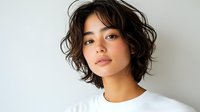 Young Asian woman with short wavy dark hair and natural makeup wearing white t-shirt against light background, looking directly at camera with confident expression.