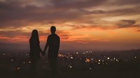 Silhouetted couple holding hands against dramatic golden sunset sky with city lights below, romantic evening scene for relationships and love concepts.