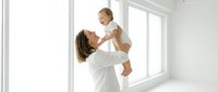 Young Caucasian mother in white outfit lifting laughing baby in bright minimal room, sharing joyful bonding moment in natural light.
