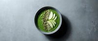 Green smoothie bowl with sliced cucumber, hemp seeds, and coconut flakes in black bowl on gray surface, healthy breakfast and nutrition concept.