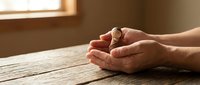 Cupped hands in warm sunlight on wooden table suggesting prayer, meditation, hope, protection, and spiritual contemplation in peaceful indoor setting.
