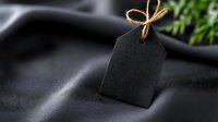 Black gift tag with rustic twine bow on dark fabric background with soft green foliage, creating minimalist composition for special occasions and celebrations.