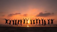 Silhouettes of people jumping with raised arms on beach at sunset, celebrating freedom and joy against vibrant orange sky.