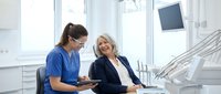 Smiling senior Caucasian woman in dental chair consulting with female dentist in scrubs holding tablet in modern clinic.