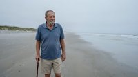 Senior man with gray beard wearing casual blue shirt standing on empty sandy beach holding walking stick during overcast day, suitable for lifestyle, travel, and wellness content.