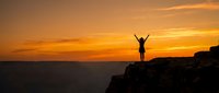 Woman hiker celebrating success on mountain cliff edge during golden sunset with arms raised in victory pose against dramatic orange sky and ocean horizon.